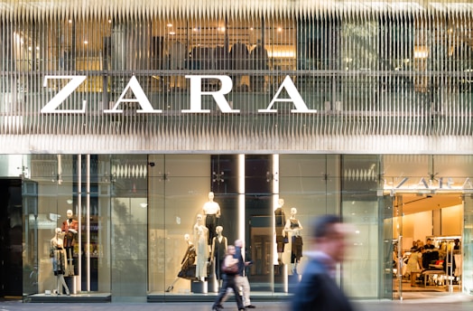 A modern retail store front with large glass windows displaying mannequins dressed in fashionable clothing. The large, bold signage above spells out the store's name. There are people walking past, slightly blurred, indicating motion and a bustling city environment.