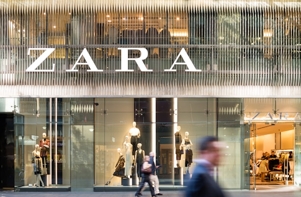 A modern retail store front with large glass windows displaying mannequins dressed in fashionable clothing. The large, bold signage above spells out the store's name. There are people walking past, slightly blurred, indicating motion and a bustling city environment.