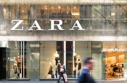 A modern retail store front with large glass windows displaying mannequins dressed in fashionable clothing. The large, bold signage above spells out the store's name. There are people walking past, slightly blurred, indicating motion and a bustling city environment.