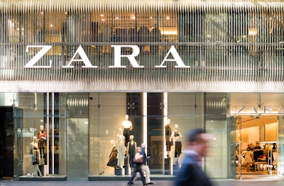 A modern retail store front with large glass windows displaying mannequins dressed in fashionable clothing. The large, bold signage above spells out the store's name. There are people walking past, slightly blurred, indicating motion and a bustling city environment.