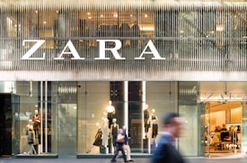 A modern retail store front with large glass windows displaying mannequins dressed in fashionable clothing. The large, bold signage above spells out the store's name. There are people walking past, slightly blurred, indicating motion and a bustling city environment.