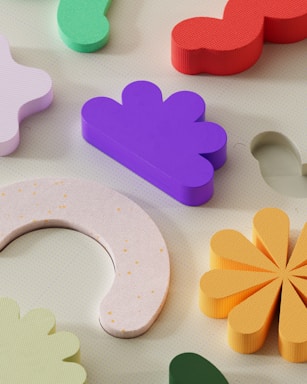 Colorful and textured geometric shapes arranged on a light-colored surface. The shapes include a purple cloud-like form, a red wavy piece, a green abstract shape, a crescent with tiny yellow dots, and a daisy-like yellow design.