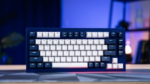 A mechanical keyboard with navy blue and white keycaps is placed on a desk with a geometric-patterned mat. The background features a blurred indoor setting with soft lighting, possibly a lamp and some furniture.