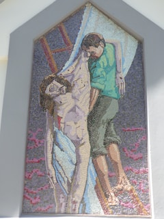 A mosaic artwork depicting a somber scene with two figures, one of whom appears lifeless, being held by another person. The figures are composed of small colored tiles, with one person wearing a greenish garment and the other depicted with noticeable wounds.
