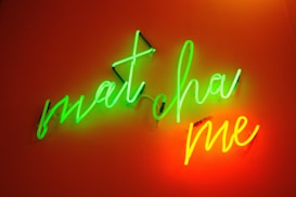 Bright neon text spelling 'matcha me' in a cursive style. The words are illuminated in green and orange neon lights against a warm, reddish-brown background.
