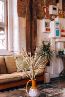 A cozy interior setting featuring a brown sofa with a green cushion in the background. In the foreground, there is a ceramic vase with an earthy tone, containing tall dried flowers or branches. The walls have a rustic appearance with a mix of exposed brick and wooden elements. Various colorful notices and a plant on a stand add detail to the background.