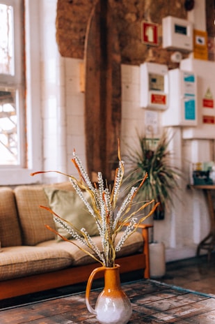 A cozy interior setting featuring a brown sofa with a green cushion in the background. In the foreground, there is a ceramic vase with an earthy tone, containing tall dried flowers or branches. The walls have a rustic appearance with a mix of exposed brick and wooden elements. Various colorful notices and a plant on a stand add detail to the background.