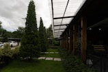 A well-maintained landscaped area with neatly trimmed trees and shrubs is bordered by a modern structure featuring a transparent corrugated roof supported by wooden beams. Cars are parked nearby, partially visible on the left side. The sky is overcast, adding depth to the natural setting.