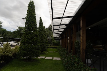 A well-maintained landscaped area with neatly trimmed trees and shrubs is bordered by a modern structure featuring a transparent corrugated roof supported by wooden beams. Cars are parked nearby, partially visible on the left side. The sky is overcast, adding depth to the natural setting.