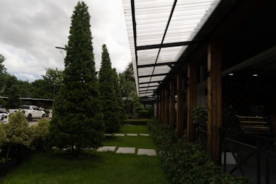 A well-maintained landscaped area with neatly trimmed trees and shrubs is bordered by a modern structure featuring a transparent corrugated roof supported by wooden beams. Cars are parked nearby, partially visible on the left side. The sky is overcast, adding depth to the natural setting.
