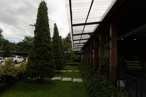 A well-maintained landscaped area with neatly trimmed trees and shrubs is bordered by a modern structure featuring a transparent corrugated roof supported by wooden beams. Cars are parked nearby, partially visible on the left side. The sky is overcast, adding depth to the natural setting.