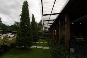 A well-maintained landscaped area with neatly trimmed trees and shrubs is bordered by a modern structure featuring a transparent corrugated roof supported by wooden beams. Cars are parked nearby, partially visible on the left side. The sky is overcast, adding depth to the natural setting.
