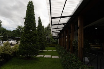 A well-maintained landscaped area with neatly trimmed trees and shrubs is bordered by a modern structure featuring a transparent corrugated roof supported by wooden beams. Cars are parked nearby, partially visible on the left side. The sky is overcast, adding depth to the natural setting.