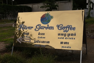 A large, golden-colored outdoor sign with blue text advertising 'River Garden Coffee.' The sign includes English and Khmer language offerings such as coffee, cakes, cold drinks, milk tea, and meals. It is set against a backdrop of grass and a concrete path.