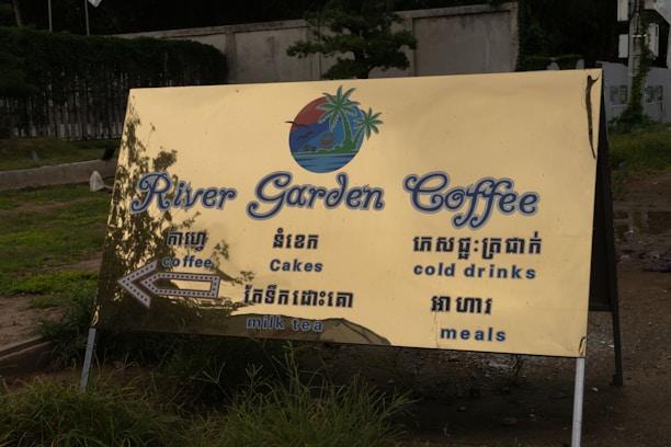 A large, golden-colored outdoor sign with blue text advertising 'River Garden Coffee.' The sign includes English and Khmer language offerings such as coffee, cakes, cold drinks, milk tea, and meals. It is set against a backdrop of grass and a concrete path.