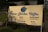 A large, golden-colored outdoor sign with blue text advertising 'River Garden Coffee.' The sign includes English and Khmer language offerings such as coffee, cakes, cold drinks, milk tea, and meals. It is set against a backdrop of grass and a concrete path.