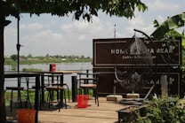 A serene outdoor seating area with a view of a calm body of water in the background. The space features a wooden deck with multiple tables and chairs arranged neatly. A cozy egg-shaped swing chair is positioned to the right, and there is lush greenery surrounding the area, including overhanging trees and banana plants. A sign in the background suggests this is a cafe or restaurant setting.