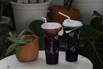 On a white table, two tall cups with intricate leaf designs and straws stand next to a small potted succulent plant. The background features large green leaves and another plant pot, creating a lush, natural setting.