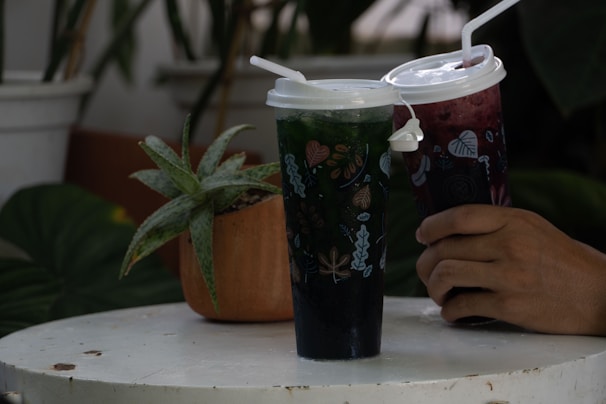 Two plastic cups with decorative leaf designs contain beverages, one of which is green and the other dark red. A person is holding the red drink. The cups have white lids and straws. Nearby, a potted succulent plant sits on a round white table, surrounded by green foliage.