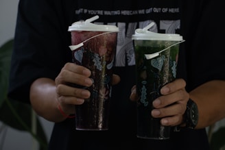 A person is holding two large, lidded plastic cups with straws, one filled with a red drink and the other with a green drink. The cups feature decorative designs with leaf patterns. The person is wearing a dark shirt and a watch on their left wrist.