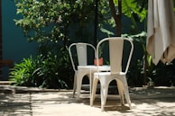In a shaded outdoor setting, two white metal chairs are positioned around a small table topped with a potted plant. The area is surrounded by lush green foliage, under natural sunlight that casts soft shadows on the ground.