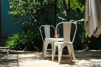 In a shaded outdoor setting, two white metal chairs are positioned around a small table topped with a potted plant. The area is surrounded by lush green foliage, under natural sunlight that casts soft shadows on the ground.