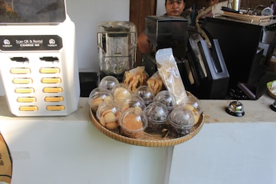 A cafe counter displaying various food items under plastic domes, a charging station labeled 'Scan QR & Rental Charge Me,' a blender, and coffee-related equipment. A person stands behind the counter.