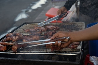 A person is grilling meat on an outdoor barbecue grill. The grill is covered with a variety of meat pieces, which appear to be seasoned and partially cooked. The hands of the person are visible, using tongs to turn or rearrange the meat.