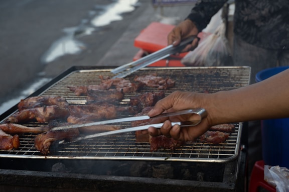 A person is grilling meat on an outdoor barbecue grill. The grill is covered with a variety of meat pieces, which appear to be seasoned and partially cooked. The hands of the person are visible, using tongs to turn or rearrange the meat.