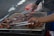 A person is grilling meat on an outdoor barbecue grill. The grill is covered with a variety of meat pieces, which appear to be seasoned and partially cooked. The hands of the person are visible, using tongs to turn or rearrange the meat.