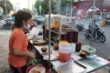 A woman is working at a busy street food stall. She is wearing a mask and gloves while handling food and packing it in a container. The stall has various food items, including cooked meat and vegetables, displayed on metal shelves. There are numerous bowls, containers, and ingredients placed on the counter. The background shows a bustling street with motorcycles and cars driving by, and pedestrians walking.