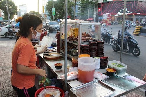 A woman is working at a busy street food stall. She is wearing a mask and gloves while handling food and packing it in a container. The stall has various food items, including cooked meat and vegetables, displayed on metal shelves. There are numerous bowls, containers, and ingredients placed on the counter. The background shows a bustling street with motorcycles and cars driving by, and pedestrians walking.