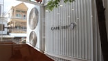 The image features the interior of a modern cafe with large glass windows, allowing a view of the buildings outside. There is a sign on the wall that reads 'Café 101' next to an illustration of a steaming coffee cup. Two large, decorative umbrellas are mounted on the wall next to the windows. Part of a tree and some orange-brown furniture are visible inside the cafe.