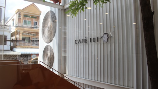 The image features the interior of a modern cafe with large glass windows, allowing a view of the buildings outside. There is a sign on the wall that reads 'Café 101' next to an illustration of a steaming coffee cup. Two large, decorative umbrellas are mounted on the wall next to the windows. Part of a tree and some orange-brown furniture are visible inside the cafe.