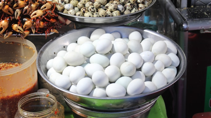 A market scene with various foods displayed in metal bowls and containers. The main focus is on a large bowl of white eggs. Above it, smaller quail eggs are placed in a dish. To the side, a collection of small crabs with orange claws is skewered on sticks. Containers filled with what appears to be sauces or spices are also visible.
