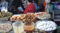 A vibrant food market stall displaying a variety of foods including cooked crabs, quail eggs, and snails. Large containers filled with white eggs, small seashells, and several jars containing sauces or condiments are also visible. People are present, preparing or purchasing food, creating a lively and bustling atmosphere.