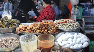 A vibrant food market stall displaying a variety of foods including cooked crabs, quail eggs, and snails. Large containers filled with white eggs, small seashells, and several jars containing sauces or condiments are also visible. People are present, preparing or purchasing food, creating a lively and bustling atmosphere.