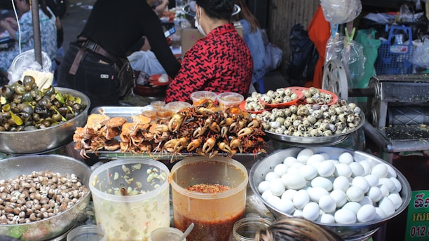 A vibrant food market stall displaying a variety of foods including cooked crabs, quail eggs, and snails. Large containers filled with white eggs, small seashells, and several jars containing sauces or condiments are also visible. People are present, preparing or purchasing food, creating a lively and bustling atmosphere.