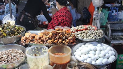 A vibrant food market stall displaying a variety of foods including cooked crabs, quail eggs, and snails. Large containers filled with white eggs, small seashells, and several jars containing sauces or condiments are also visible. People are present, preparing or purchasing food, creating a lively and bustling atmosphere.