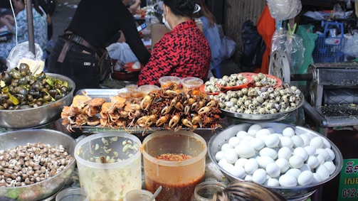 A vibrant food market stall displaying a variety of foods including cooked crabs, quail eggs, and snails. Large containers filled with white eggs, small seashells, and several jars containing sauces or condiments are also visible. People are present, preparing or purchasing food, creating a lively and bustling atmosphere.