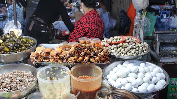 A vibrant food market stall displaying a variety of foods including cooked crabs, quail eggs, and snails. Large containers filled with white eggs, small seashells, and several jars containing sauces or condiments are also visible. People are present, preparing or purchasing food, creating a lively and bustling atmosphere.