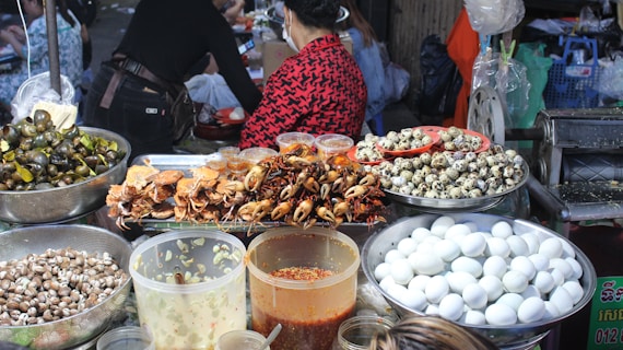 A vibrant food market stall displaying a variety of foods including cooked crabs, quail eggs, and snails. Large containers filled with white eggs, small seashells, and several jars containing sauces or condiments are also visible. People are present, preparing or purchasing food, creating a lively and bustling atmosphere.