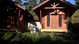 Two wooden cabins with pointed roofs are situated side by side in a lush, green area. The cabins are surrounded by plants and small trees, and a concrete walkway leads up to them. Sunlight casts dramatic shadows on the structures, highlighting their wooden textures. One cabin has a decorative object or sign in front, and the surrounding greenery adds a serene, natural atmosphere.