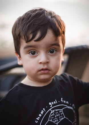 A young child with dark hair and big eyes gazes directly at the camera. The child is wearing a black shirt with white text and graphic design. The background is blurred, suggesting an outdoor setting in soft light.