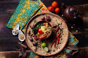 A vibrant and artistic food presentation featuring a bowl of noodles garnished with boiled egg halves, red chili peppers, tomatoes, herbs, and spices. The food is placed on a woven bamboo tray surrounded by additional ingredients like chili peppers, onions, and lime. The setting includes a teapot and is adorned with colorful fabric in an ethnic pattern.