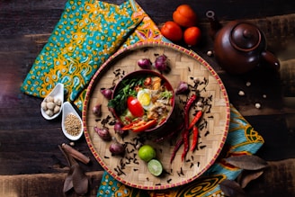 A vibrant and artistic food presentation featuring a bowl of noodles garnished with boiled egg halves, red chili peppers, tomatoes, herbs, and spices. The food is placed on a woven bamboo tray surrounded by additional ingredients like chili peppers, onions, and lime. The setting includes a teapot and is adorned with colorful fabric in an ethnic pattern.