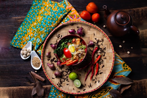 A vibrant and artistic food presentation featuring a bowl of noodles garnished with boiled egg halves, red chili peppers, tomatoes, herbs, and spices. The food is placed on a woven bamboo tray surrounded by additional ingredients like chili peppers, onions, and lime. The setting includes a teapot and is adorned with colorful fabric in an ethnic pattern.
