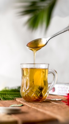 A spoon drizzles golden liquid, likely honey, into a glass mug filled with a warm, amber-colored beverage. The scene is set against a soft, blurred background with hints of greenery, suggesting a natural or calming environment.