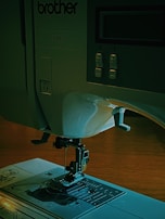 A sewing machine with a visible needle and presser foot on a flat surface. The control panel includes buttons and a display screen. The model number CE8080 is visible, and the brand name 'brother' is marked on the machine.