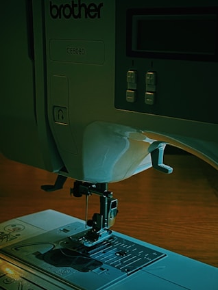 A sewing machine with a visible needle and presser foot on a flat surface. The control panel includes buttons and a display screen. The model number CE8080 is visible, and the brand name 'brother' is marked on the machine.